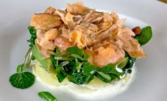 A plated dish featuring flaked salmon on a bed of fresh greens and sliced potatoes, served atop a layer of creamy white sauce on a white plate.