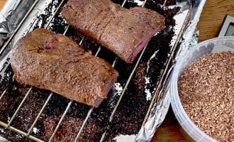 Two pieces of cooked meat rest on a metal rack inside a foil-lined roasting tin. Beside the tin is a plastic container filled with wood chips, likely for smoking the meat.