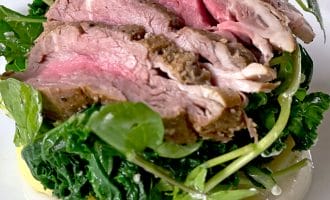 Slices of medium-rare roast beef are served on a bed of leafy greens and watercress atop sliced potatoes with a creamy white sauce, arranged on a white plate.