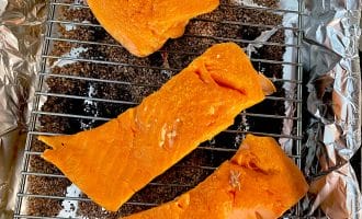 Three pieces of raw salmon fillet are placed on a metal rack over a layer of brown wood chips on aluminium foil, ready to be smoked or cooked.