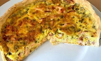A baked quiche with a golden crust and a savoury filling, featuring visible pieces of vegetables and meat, sits on a white plate with a large slice removed.