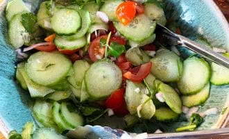 A bowl of fresh cucumber salad with sliced cucumbers, cherry tomatoes, onions, and coriander, served with metal tongs on a wooden table.
