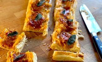 Rectangular puff pastry tart topped with orange mashed squash, crispy sage leaves, and slices of cured meat, cut into pieces on a wooden surface next to a knife.