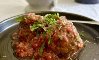 Two meatballs topped with tomato sauce and chopped fresh herbs are served on a dark plate. Some grated cheese is visible on top and around the plate, with a spoon resting beside the meatballs.