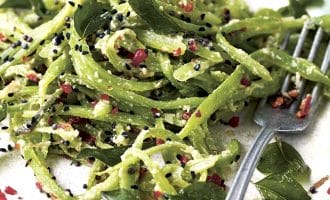 A plate of green pasta topped with black seeds, red chilli flakes, grated cheese, and fresh herbs. A fork rests on the plate beside the pasta.