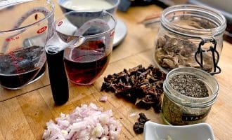 Chopped shallots, minced garlic, dried mushrooms, dried herbs, red wine, and cream are arranged on a wooden kitchen worktop, ready for cooking. Measuring jugs and glass jars hold some of the ingredients.