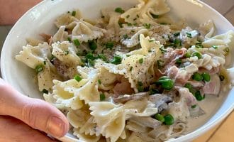A person holds a white bowl filled with farfalle pasta in a creamy sauce, mixed with peas, ham, mushrooms, and garnished with chopped chives and ground black pepper.