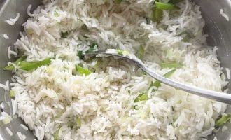 A pot of cooked white basmati rice mixed with chopped spring onions, with a metal spoon resting inside the pot.