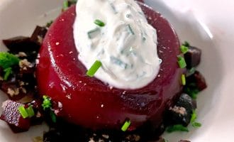 A round serving of beet jelly topped with a dollop of herbed cream sits on a plate, surrounded by small diced beetroot and sprinkled with fresh chives.