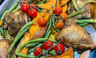 Baked chicken thighs, sweet potato wedges, green beans, and vine tomatoes arranged on a metal tray, seasoned and ready to eat.