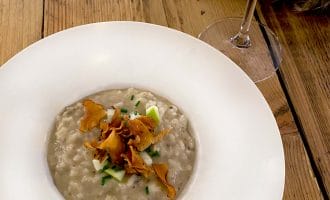 A white bowl of creamy risotto topped with crispy crisps and chopped herbs sits on a wooden table next to a glass of rosé wine.