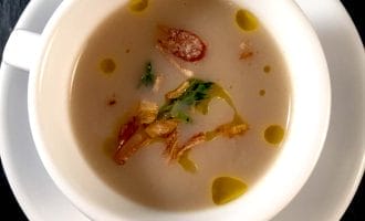A white cup on a white saucer filled with creamy soup, garnished with crispy fried shallots, green herbs, and drops of oil, set on a dark surface.