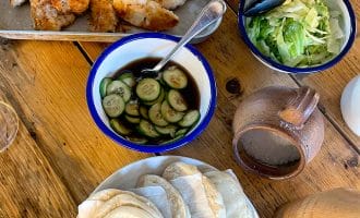 A wooden table set with a tray of breaded chicken goujons, a bowl of sliced cucumbers in sauce, a bowl of shredded lettuce, a pile of folded flatbreads, and a brown pottery jug.