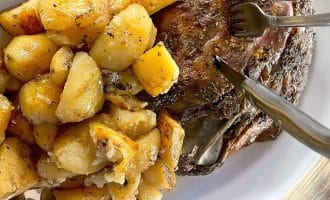 A white plate holding roast potatoes seasoned with herbs and a piece of roast meat with a fork and knife cutting into it, on a wooden table.