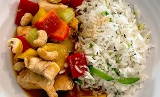 A white bowl filled with white rice mixed with vegetables, served alongside stir-fried chicken with red and yellow peppers, celery, pineapple, and topped with cashew nuts in a savoury sauce.