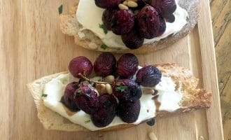 Three slices of crusty bread topped with creamy cheese, roasted grapes, pine nuts, and fresh herbs are arranged on a wooden serving board.