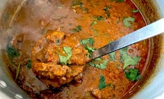A pot of rich, red-brown curry garnished with fresh coriander, with a ladle lifting a portion of the meat and sauce. Steam rises from the hot dish, indicating it is freshly cooked.