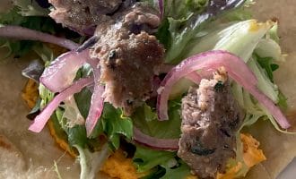 A close-up of a flatbread topped with mixed leaves, sliced red onions, grilled meat pieces, and an orange spread, ready to be wrapped.