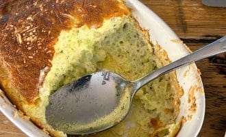A close-up of a baked courgette soufflé in a white oval dish, with a spoon resting inside and part of the soufflé scooped out, showing its fluffy, airy texture. The dish sits on a rustic wooden table.
