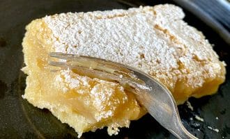 A slice of lemon bar covered with icing sugar sits on a dark plate, with a fork cutting into its soft, creamy texture.