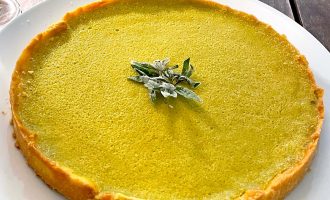 A round matcha green tea tart with a golden crust, topped with a small cluster of sugared mint leaves, sits on a white plate on a wooden table.