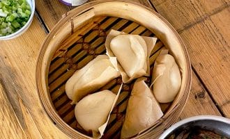 A bamboo steamer with five folded bao buns sits on a wooden table, surrounded by bowls of spring onions, herbs, a jar of sauce, tongs, and a pot of shredded meat.