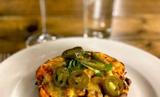 A baked sweet potato half topped with melted cheese, beans, and sliced jalapeños sits on a white plate. In the background are a glass of red drink and a tall glass of water on a wooden table.