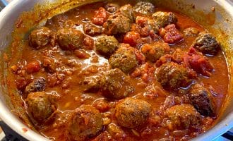 A frying pan filled with meatballs simmering in a rich tomato sauce with chunks of tomatoes, set on a hob with tiled splashback in the background.