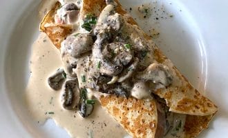 A savoury crêpe filled with mushrooms and topped with creamy mushroom sauce and chopped herbs, served on a white plate.