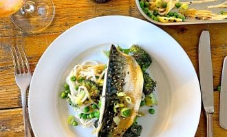 A white plate with grilled fish on top of noodles, broccoli, peas, and bean sprouts. A fork and knife are on either side, with a glass of drink and a bottle of soy sauce on a wooden table.