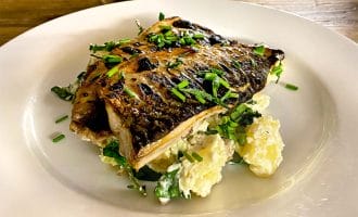 Grilled fish fillet garnished with chopped chives, served on a bed of salad leaves and creamy potato salad, presented on a white plate atop a wooden table.