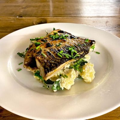 Grilled fish fillet garnished with chopped chives, served on a bed of salad leaves and creamy potato salad, presented on a white plate atop a wooden table.