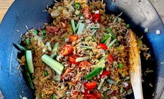 A wok filled with fried rice mixed with peas, tomatoes, cucumber slices, greens, and minced meat, with a wooden spoon resting on the side. The dish sits on a wooden table.