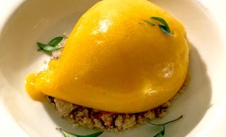 A bright yellow scoop of mango sorbet sits on a bed of crumbled biscuit, garnished with a few small green herb leaves, served in a white bowl.