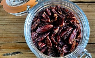 A glass jar filled with roasted pecan halves sits open on a rustic wooden surface, with the orange-rimmed lid resting to the side and attached by a metal clasp.