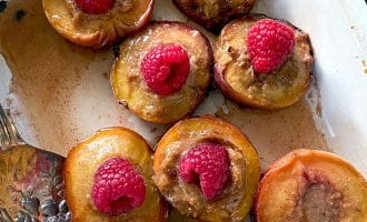 Halved baked peaches topped with a dollop of brown filling and fresh raspberries, arranged in a white baking dish with a serving spoon at the side.