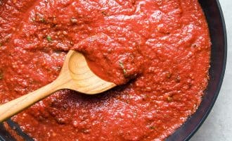 A wooden spoon stirs a thick, red tomato sauce with herbs in a black pan, viewed from above.