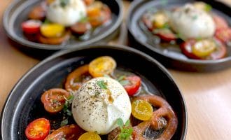 Five plates each feature a salad of sliced red and yellow heritage tomatoes, topped with a ball of burrata cheese, fresh herbs, black pepper, and a drizzle of olive oil, arranged on dark round plates.