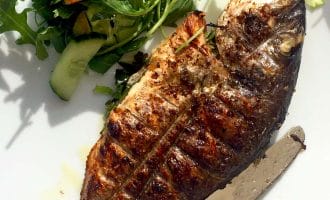 Grilled whole fish served on a white plate with a side of fresh green salad, including rocket and cucumber, accompanied by a fork and a knife.
