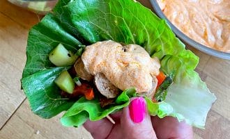 A hand with pink nail varnish holds a lettuce wrap filled with grilled chicken, diced vegetables, and topped with a dollop of orange creamy sauce. Bowls of salad and sauce are in the background on a wooden surface.