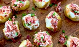 Bite-sized baked potato pieces topped with soured cream, crumbled bacon, and chopped chives are arranged on a wooden surface.