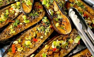 Roasted aubergine halves topped with chopped spring onions, red chilli, sesame seeds, and herbs are arranged on a tray lined with foil. Metal tongs are placed on the right side of the tray.