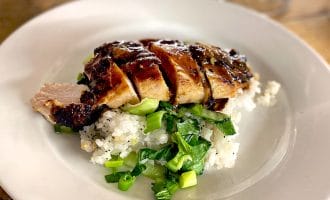 Sliced grilled chicken breast served on a bed of white rice and chopped green vegetables, arranged on a white plate on a wooden table. A blurred wine glass is visible in the background.
