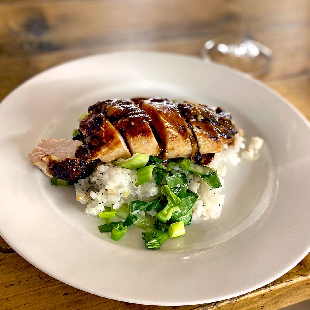 Sliced grilled chicken breast served on a bed of white rice and chopped green vegetables, arranged on a white plate on a wooden table. A blurred wine glass is visible in the background.