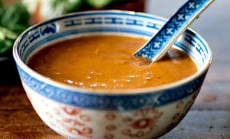 A blue-and-white patterned bowl filled with brown peanut sauce, with a matching spoon resting inside. The bowl sits on a wooden surface, and green herbs are blurred in the background.