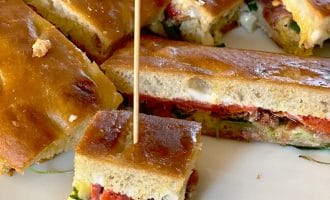 A close-up of sliced focaccia sandwiches with fillings such as vegetables and cheese, served on a white plate. One small sandwich piece is secured with a cocktail stick.