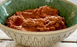 A decorative ceramic bowl filled with a chunky, reddish-brown dip sits on a wooden table, with a blurred outdoor scene in the background.