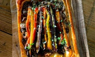 A rectangular vegetable tart with flaky golden pastry, topped with colourful roasted carrots, herbs, and a dark spread, sits on baking parchment on a wooden chopping board in sunlight.