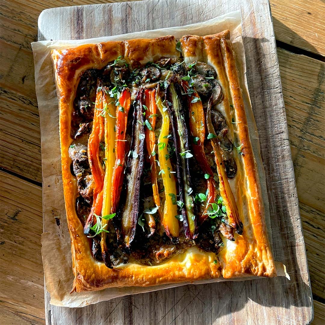 A rectangular vegetable tart with flaky golden pastry, topped with colourful roasted carrots, herbs, and a dark spread, sits on baking parchment on a wooden chopping board in sunlight.