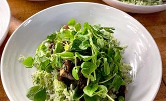 A white bowl filled with green herb rice, topped with sautéed mushrooms and fresh leafy microgreens, sits on a wooden table with similar bowls in the background.